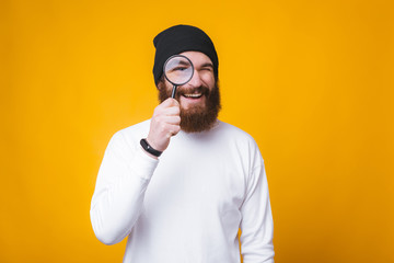 Young bearded man is looking through a magnifying glass and smiling near yellow wall.