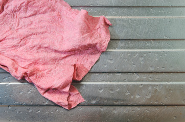 Single wet dirty used pink dishcloth (washcloth). Placed on top of a steel kitchen sink with water droplets all around. No people, real raw shot. Copy space. Concept for unhealthy and germs growth