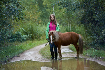 Teenage girl stands with a little horse on the dirt road with big puddles
