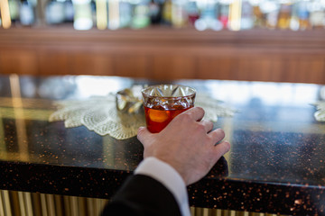 Rich and successful businessman holds a glass of Scotch whisky with an ice cube on a wooden table, sitting at the bar . Hand holding liquor whiskey or cognac in the glass on the background of the bar