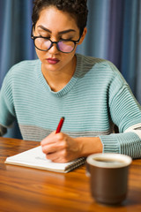 A woman wearing glasses sits at a table drinking coffee and writing in a journal