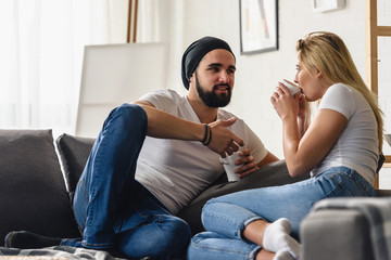 Young hipster couple sitting on sofa at home talking and drinking coffee