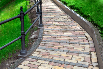 pedestrian walkway made of stone tiles and a granite curb with iron railings in the park with a rise up the slope.