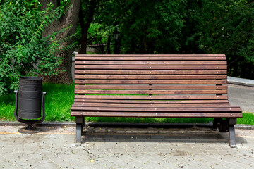 a wooden bench made of brown boards on the back and an iron black litter bin on a stone sidewalk in...