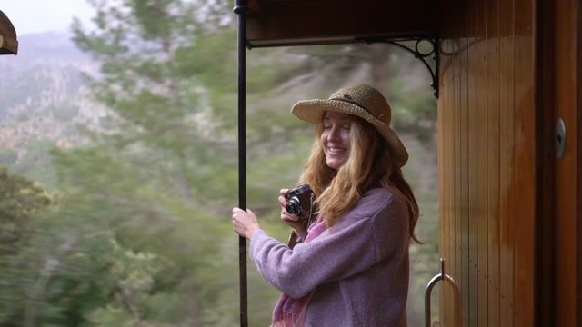 A young woman in a straw hat enjoying traveling on an old train, taking pictures of beautiful tourist locations using vintage camera, feeling excited and happy