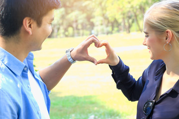 Sweet couple showing love each other in park, young beautiful lover making heart shape while spending time together and having romantic moment in summer garden.