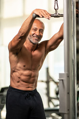 Obraz premium Muscular shirtless mature older bodybuilding athlete with balding gray hair performing a back exercise on a machine in a gym smiling at the camera