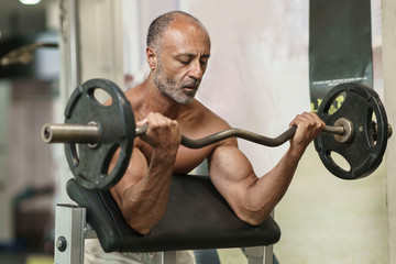 Muscular shirtless mature older bodybuilding athlete with balding gray hair does a bicep curl in a...