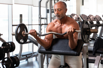 Muscular shirtless mature older bodybuilding athlete with balding gray hair does a bicep curl in a...