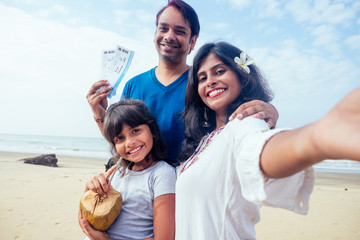 Young indian family relaxing and taking selfie on camera smartphone with ticket and coconut in Goa