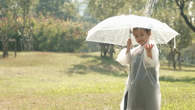 Little girl wear raincoat and transparent umbrella play with artificial rain in the garden with morning light.