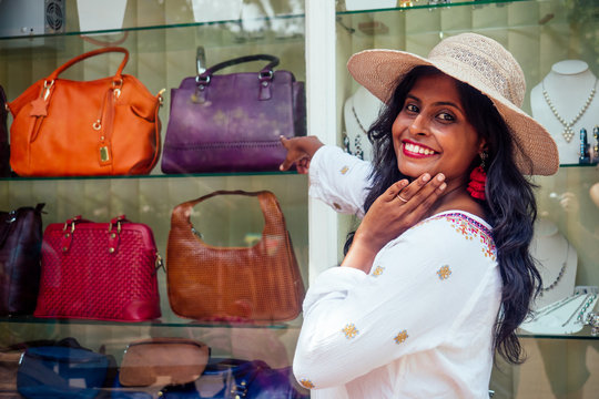 happy indian woman wearing straw hat and standind near showcase with fashion bags in India Goa market