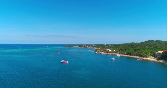 Aerial forward over blue ocean and boats, along island coast, Roatan