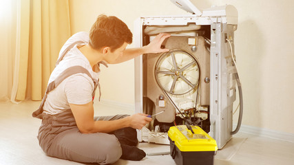 professional master in grey jumpsuit pants opens back panel of broken washing machine top loader, sunlight