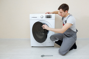 professional worker in grey uniform checking broken modern washing machine on wooden floor near...