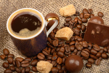 A Cup of coffee, coffee beans, pieces of brown sugar and chocolate on a background of rough homespun fabric. Close up.