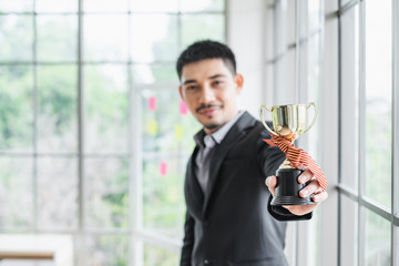 An Asian business man in suit holding a trophy in front of bright window shallow focus on trophy.