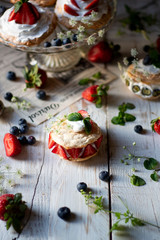 Mini pavlovas on a cake holder and on white wooden table, sprinkled with fresh strawberries and blueberries, mint leafs and flowers