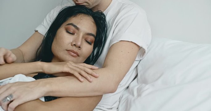 Close up of lover sitting in bed thinking about future. Caucasian male with his girlfriend, asian woman in black lingerie. Happy lover in bed concept.