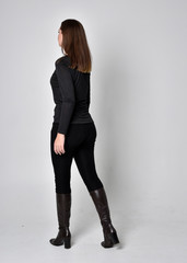 full length portrait of a pretty brunette girl wearing a black shirt leather boots. Standing pose, facing away from the camera,  on a grey studio background.