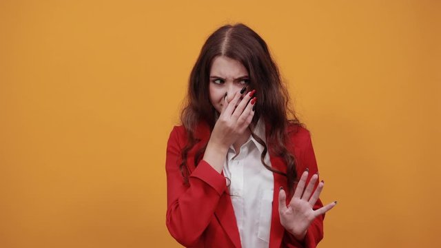 Unhappy young woman in fashion white shirt and red jacket covers nose from bad smell isolated on orange background in studio. People sincere emotions, lifestyle concept.