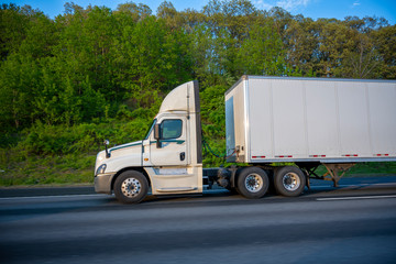 Profile of white big rig day cab semi truck with roof spoiler transporting commercial cargo in dry van semi trailer going on the wide highway with green trees on the roadside