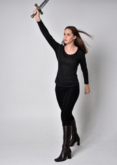 full length portrait of a pretty brunette girl wearing a black shirt and leather boots, holding a sword. Standing pose, holding a sword, on a grey studio background.