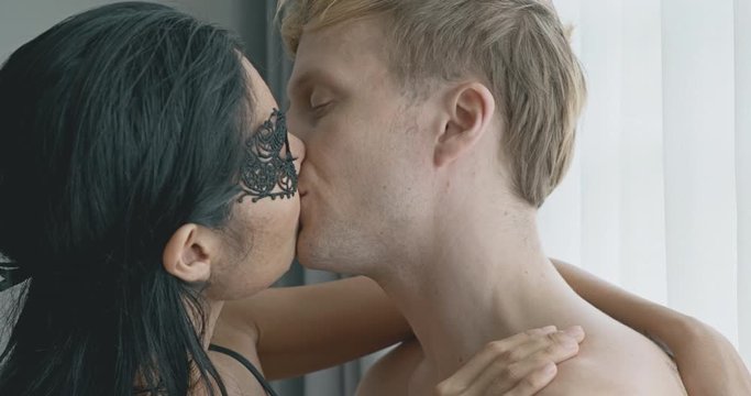Close up of lover kissing passionately, standing up. Caucasian male with his girlfriend in black mask, asian woman in black lingerie. Safe sex and bdsm roleplay concept.