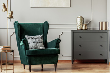 Emerald green wing back chair with pillow in grey living room interior with wooden commode - obrazy, fototapety, plakaty