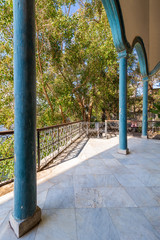 Elements of covered terrace with blue stone pillars, and marble floor fenced with metal forged fence and surrounded with green trees in sunny summer day
