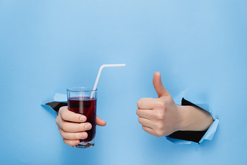 Female hand holding a glass of fresh red juice with a straw through torn blue paper wall. Fruit...