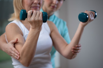 Fototapeta na wymiar Professional physiotherapist helping senior woman to lift hand weights