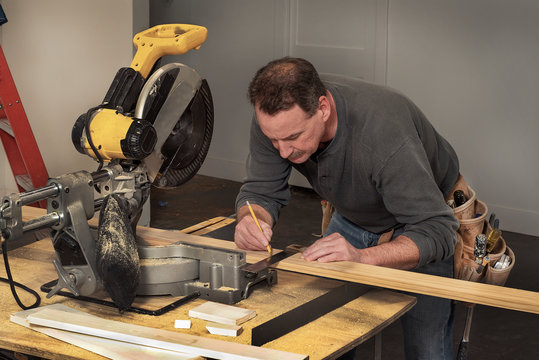 Male Carpenter Wearing Old Tool Belt, Marking Wood Board With Old Square And Pencil, Next To Electric Chop Saw, At Fixer Upper Home Residential Construction Site