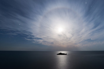 Full moon halo in the starry sky above the sea with a small rock