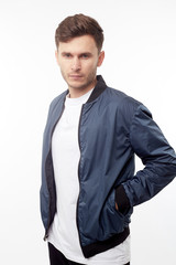 Young european man in white sweater and black pants, blue bomber jacket posing on white background....