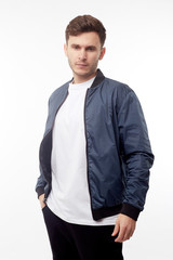 Young european man in white sweater and black pants, blue bomber jacket posing on white background....