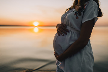 Pregnancy. A pregnant girl in a white dress holds on to a big belly on a background of sunset and river. Waiting for a miracle, baby. 9 months. Close-up portrait. Photography, concept.