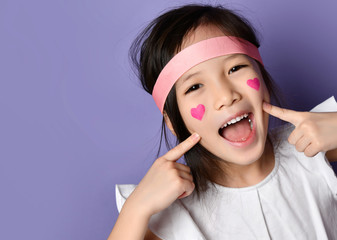 Surprised asian Korean kid girl closeup portrait Happy smiling laughing pointing fingers on cheeks with red hearts sign on cheek