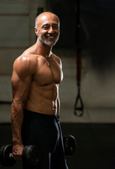 Fototapeta na wymiar Muscular shirtless mature older bodybuilding athlete with balding gray hair holding a pair of dumbbells in a gym smiling at the camera