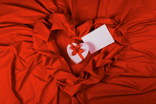 valentines greeting card. white cardboard box in shape of heart wrapped in ribbon and blank paper lay on a red cloth which repeats the form of present package. love and romance gift concept