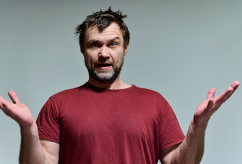 Portrait of a wild unkempt unshaven middle-aged man of 40 years in a burgundy t-shirt on a gray background. He stands right in front of the camera, talking, showing emotions. Waves his hands.