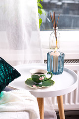 a cup of herbal tea on a table, a knitted wool blanket on a chair and candle over window . Bedroom interior