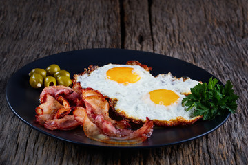 Fried eggs with bacon on a wood plate on a black background.