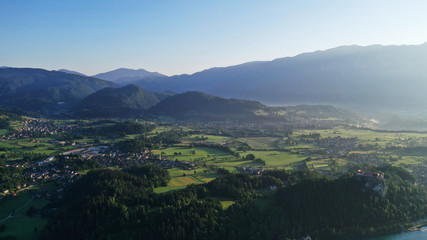 Aerial view of Bled Lake valley. Natural landscape with green meadows, fields, hills and mountains, small houses and trees. Sunny blue sky. Sun rays.