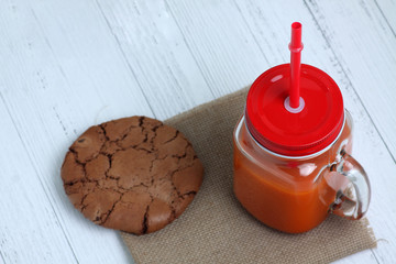 glass mug of carrot juice and a chocolate cookie on canvas napkin isolated on white wooden background flat lay