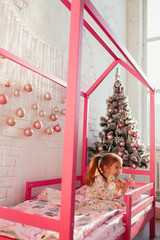 A redhead child in a pink crib-house next to a Christmas tree with toys in children's pajamas laughs and grimaces at the camera. sunlight from the window