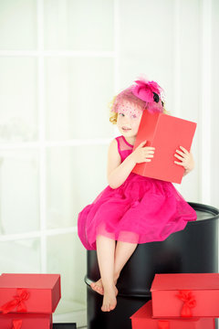Little 4 years old girl in beautiful pink dress and pink hat with veil sitting on black barrel with red gift box in hands with presents around. Birthday girl waiting for friends on birthday party.