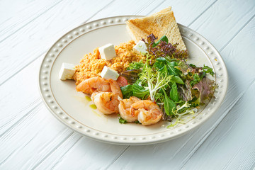 Tasty and nutritious breakfast: shrimp, couscous, feta cheese, lettuce and croutons on a white ceramic plate on a white wooden background. Healthy food. Copy space