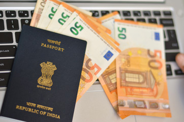 Indian Passport kept against a laptop keyboard and foreign currency Euro bills.  National emblem, Lion Capital of Ashoka from Sarnath is embossed. Concept Wealthy IT professional Berlin: Germany :2020