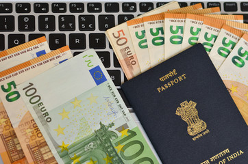Indian Passport kept against a laptop keyboard and foreign currency Euro bills.  National emblem,...
